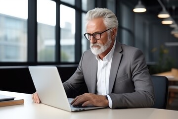 Old businessman sitting in front of a laptop in a modern office, coworking space. A gray-haired serious man works alone.