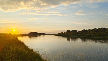 On a summer evening, the sun sets over the horizon and paints the sky in bright colors There is a ripple on the water Tall grass and deciduous trees grow along the banks and are reflected in the water