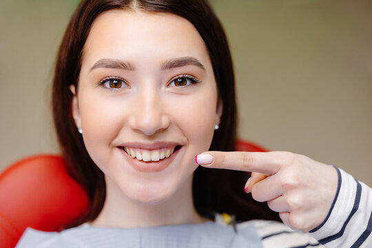 Cropped close up photo of young smiling girl, covered by napkin, pointing index finger on healthy toothy smile sitting at dental office chair. Healthcare teeth treatment