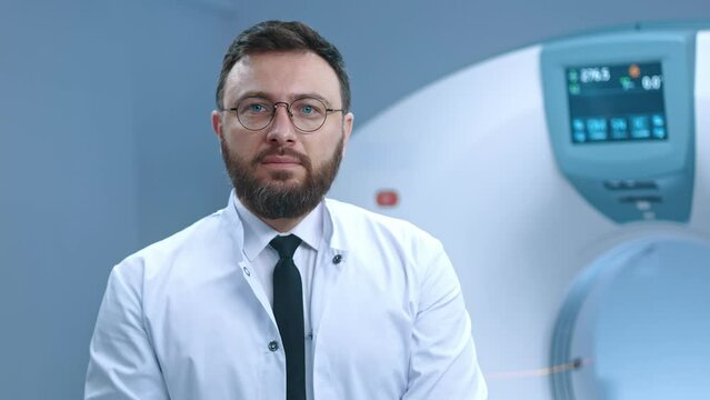 Image of responsible doctor sitting at magnetic resonance scanner. Bearded doctor in glasses. Modern equipped room for MRI scanning. Tomographer in white gown at background of MRI capsule.