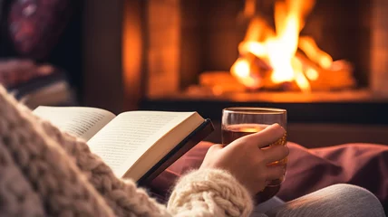 Sierkussen Woman with glass of hot tea and book near fireplace at home, closeup.   © BlazingDesigns