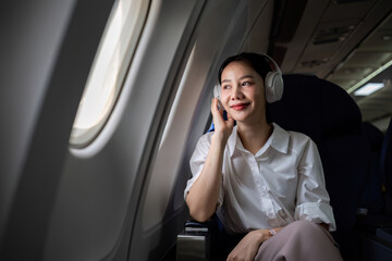 Young woman wearing headphones listening to music during travel, sitting near window in first class on airplane during flight, travel and business concept