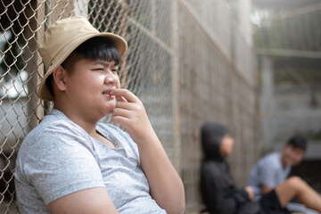 Asian teenboy in white t-shirt sits with picking teeth against a metal fence panel in a juvenile...
