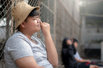 Asian teenboy in white t-shirt sits with picking mucus against a metal fence panel in a juvenile...