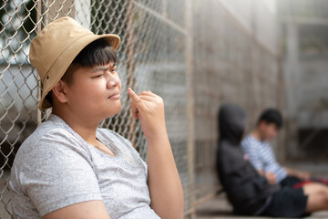 Asian teenboy in white t-shirt sits with picking mucus against a metal fence panel in a juvenile...