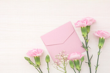 Dainty frame of pink carnations, white hazel and pink envelope on white wood grain background, with copy space