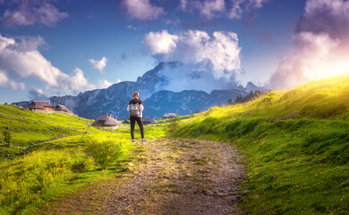 Girl on the rural road with yellow flowers and green grass in beautiful alpine mountain valley at sunset in summer. Young woman in old alpine village, blue sky with clouds. Travel and Hiking. Slovenia