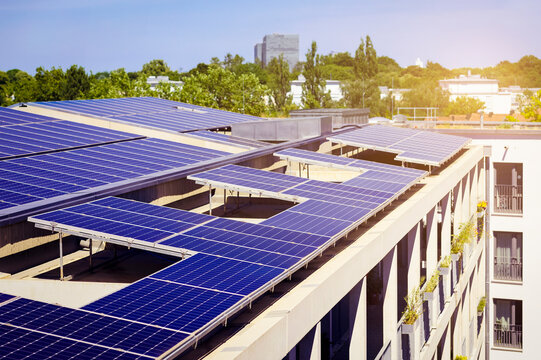 Solar Panels Roof Of Modern Multifamily High-rise Apartment Building Or Block Of Flats On Green City Backgroung With Skyscrapers And Landscaping. Solar Battery Of Residential Building. Aerial View.