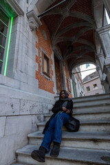 Fototapeta na wymiar This image captures a young woman in a moment of repose on the steps of a historic building. The architectural details, including the arched passageway and brickwork, provide a classic European feel