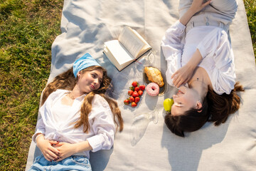 Women wearing light clothes lie on blanket. Girl friends enjoy spending time on picnic reading book and having sweets on nature