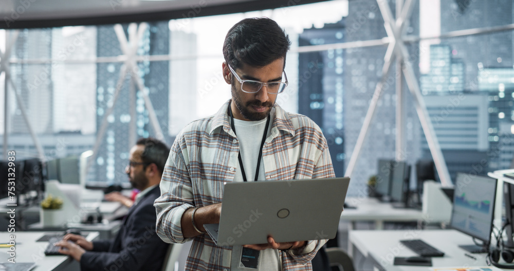 Poster Professional Indian Specialist Using Laptop while Standing in an Office Near a Window with a Big City View. Successful Software Data Engineer Optimizing Corporate Database, Update Security Network