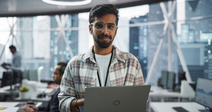 Handsome Indian Man Wearing Glasses, Charmingly Smiling and Looking at Camera. Young Intelligent Male Engineer Holding a Laptop Computer. Finance Manager Working in Modern Corporate Office
