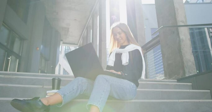 Smiling young woman student in casual clothes sitting on building's steps and working on laptop. Outdoors education and learning concept.