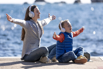 Happy child with his mother, with arms outstretched, sitting on a pier listening to music with...