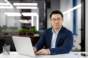 Focused Asian businessman in a suit at his desk in a contemporary office, working intently on a laptop computer.