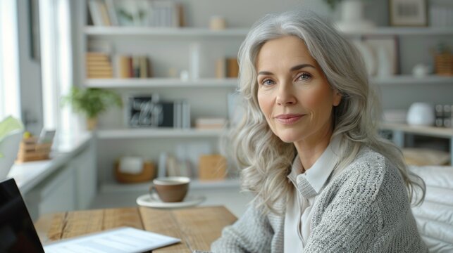 60 year old european woman, photorealistic image in close-up, with good natural lighting, in a white room with shelves, cheerful scene, at a wooden table with laptop, papers and coffee cup
