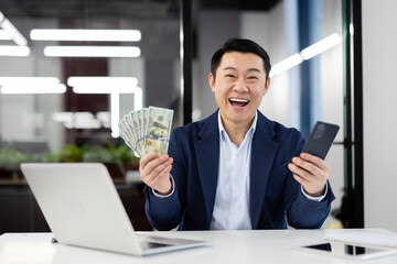 Joyful businessman in suit at office desk laughing with money in hand and a smartphone, expressing...