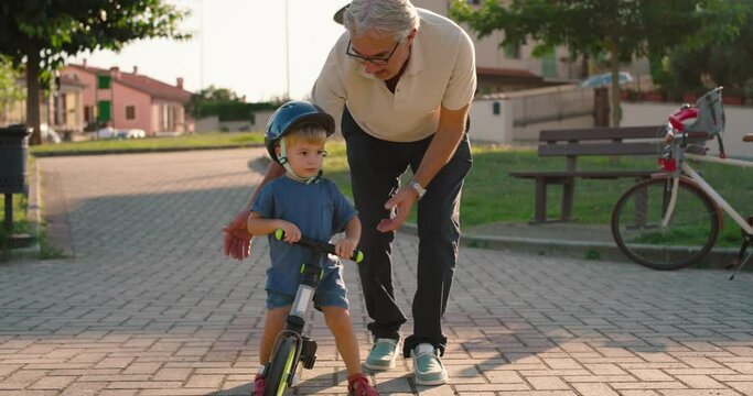 At the park, a young grandfather teaches his grandson to ride a bike, after putting a helmet on his head for safety.