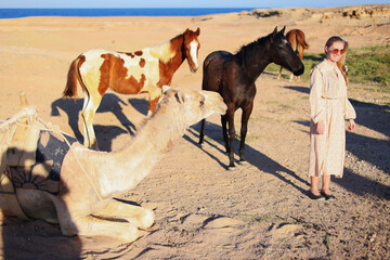 a girl in a beautiful dress, followed by two horses and a camel in the desert