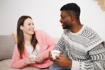 Smiling woman holding coffee while talking with african american boyfriend on couch 
