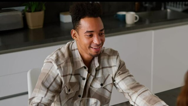 Close up of a happy young man with Black skin color in a checkered beige shirt drinks orange juice with his girlfriend during his breakfast in the kitchen