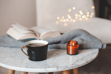 Fototapeta na wymiar Cup of coffee with open paper book and scented candle on marble table over chair and lights close up. Good morning.