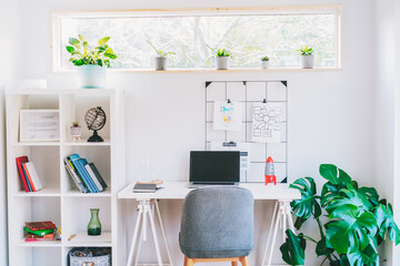 Modern cozy light workplace - white desk with laptop black screen, grid mood board with pinned notes, shelves with docs and green monstera plant at work space in home office room interior