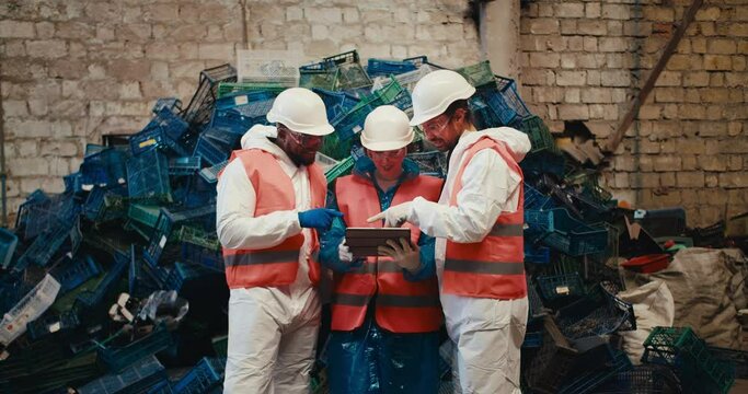 A trio of workers in white uniforms and orange vests sort out their plans on a laptop and communicate while standing near a mountain of plastic garbage at a waste recycling plant
