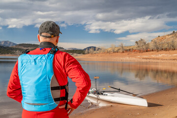 senior man wearing drysuit and life jacket with a rowing shell on lake shore, winter in northern Colorado