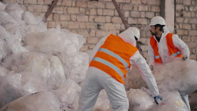 A man with Black skin in a white protective uniform an orange vest and a white helmet stacks bags of cellophane and polyethylene in one large pile together with his colleague at a large waste