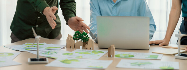 Cropped image of skilled businessman presented eco-friendly house project to general manager at meeting table with wooden block, windmill model and environmental document scatter around. Delineation.