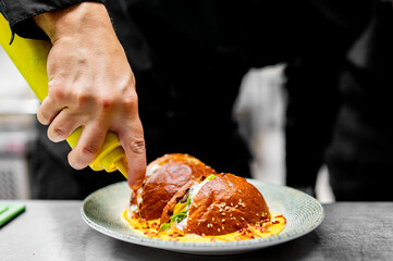 The chef’s hand, holding a yellow squeeze bottle, applies sauce onto the burger. The juicy burger rests on an elegant plate, adorned with fresh greens and sesame seeds.