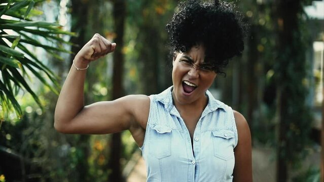 Young Confident African American Woman Flexing Arm Muscle in Green Surroundings, Strength Pose saying YEAH