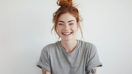 A cheerful woman with red hair tied in a bun, laughing joyfully in a casual grey t-shirt, her freckles and bright expression conveying happiness. - obrazy, fototapety, plakaty