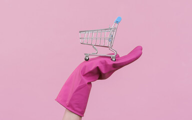Hand in purple rubber cleaning glove with mini shopping trolley on a pink background.