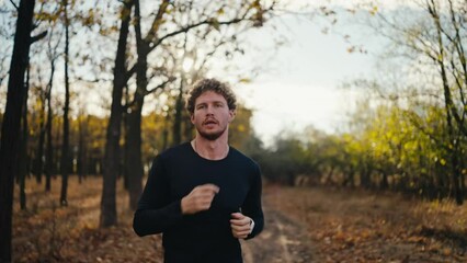 A happy confident man with curly hair and a beard in a black sports uniform with a watch on his wrist runs during his morning jog in a sunny morning autumn forest along the earth path
