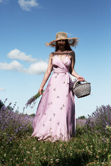 Young sexy beautiful woman in a straw hat is walking in a lavender field with a basket and a bouquet against the sky.	