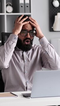 Upset Indian businessman with laptop at desk encounters a setback in the office. Expression reflect resilience and determination in the face of adversity.