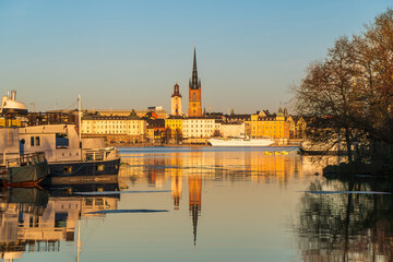 Fototapeta na wymiar Riddarholmen church reflection in the lake at sunset. Stockholm, Sweden. Blue sky, boats in foreground, late winter. Yellow buildings.