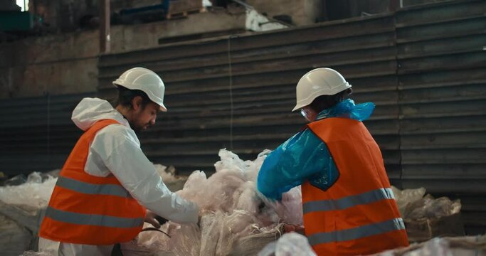 A brunette man with a beard in a white protective uniform and an orange vest together with a brunette girl presses plastic cellophane while working at a waste processing and sorting plant