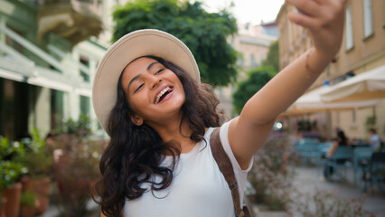 Smiling happy Indian Arabian woman female lady girl blogger influencer traveler tourist taking selfie photo on mobile phone camera travel blog for social media holidays vacation trip in city outdoors