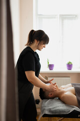 a young handsome sexy man with a naked torso undergoing a massage procedure in a sunny, bright room. The masseur presses the client's chest muscles and facial skin with his hands close-up