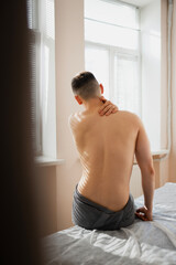 a young handsome sexy man with a naked torso undergoing a massage procedure in a sunny bright room, view from the back. A boy in a towel at a massage therapist kneads his neck with his hands