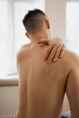 a young handsome sexy man with a naked torso undergoing a massage procedure in a sunny bright room, view from the back. A boy in a towel at a massage therapist kneads his neck with his hands