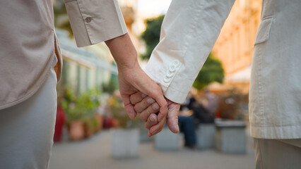 Close up back view of unrecognizable romantic senior elderly couple wife husband holding hands while walking together outside city street. Happiness people lifestyle relationship marriage resting walk