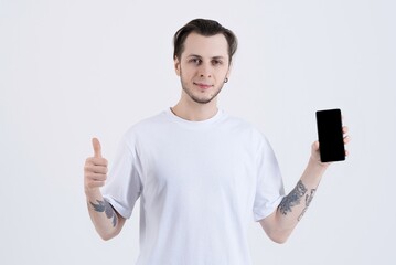 Attractive Caucasian young man with tattoos in casual clothing shows thumb up and mobile phone screen with blank copy space for advertisement isolated on white studio background.