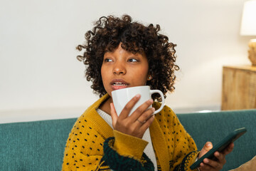 Good morning. African girl drinking coffee holding smartphone sitting on couch at home Woman with...