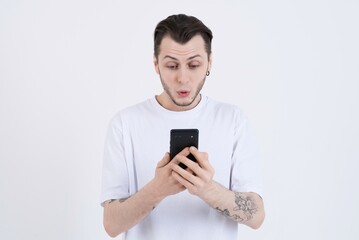 Wow! Attractive shocked caucasian young man with tattoos in casual clothes using mobile phone isolated on white studio background