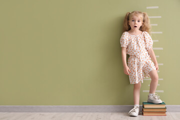 Cute little girl standing on books and measuring height near green wall