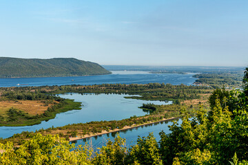 Obraz premium Zhiguli Mountains, Volga River and quiet backwater near the city of Samara, Russia. Blue water, sandy shore, tree, bush, ship. Quiet summer morning with light sky.